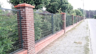 A brick and metal fence runs alongside a sidewalk lined with trees. The fence consists of alternating sections of brick pillars and metal mesh panels. Behind the fence, lush green foliage is visible, and the sidewalk is made of light gray pavers. The scene appears to be in a suburban or residential area.
