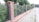 A brick and metal fence runs alongside a sidewalk lined with trees. The fence consists of alternating sections of brick pillars and metal mesh panels. Behind the fence, lush green foliage is visible, and the sidewalk is made of light gray pavers. The scene appears to be in a suburban or residential area.