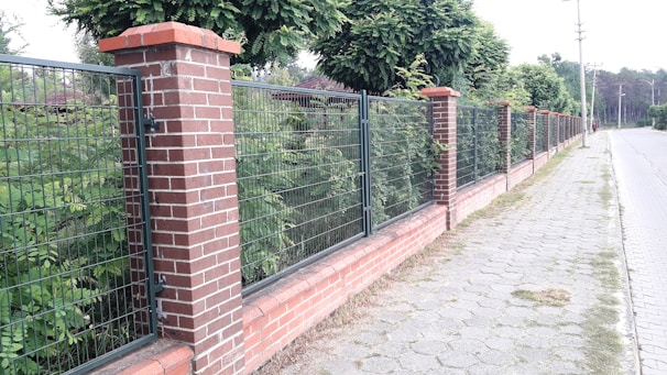 A brick and metal fence runs alongside a sidewalk lined with trees. The fence consists of alternating sections of brick pillars and metal mesh panels. Behind the fence, lush green foliage is visible, and the sidewalk is made of light gray pavers. The scene appears to be in a suburban or residential area.
