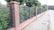 A brick and metal fence runs alongside a sidewalk lined with trees. The fence consists of alternating sections of brick pillars and metal mesh panels. Behind the fence, lush green foliage is visible, and the sidewalk is made of light gray pavers. The scene appears to be in a suburban or residential area.