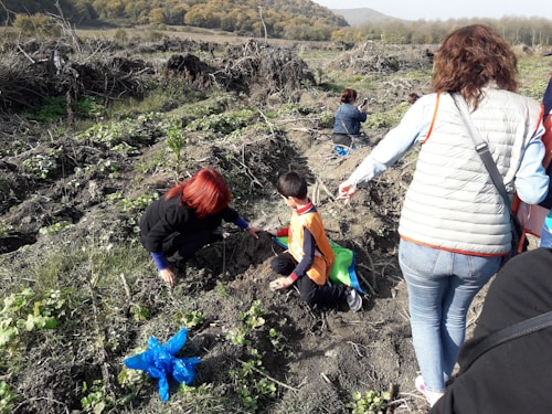 Several people are engaged in outdoor activities in a rural area with uneven, bushy terrain. A person with red hair is kneeling down near a small plant, appearing to plant or tend to it. A child wearing an orange vest is nearby, and other people are scattered in the background, some squatting or examining the ground. A blue plastic bag is visible on the ground, surrounded by green foliage under clear skies.