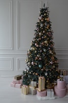 A beautifully decorated Christmas tree adorned with gold, silver, and pink baubles. There are wrapped gifts at the base of the tree in various shapes and sizes, with coordinated pink, gold, and striped wrapping. The setting is in a room with elegant white paneling on the walls.