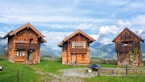 Three rustic wooden cabins are set against a backdrop of majestic mountains under a partly cloudy sky. These cabins are surrounded by lush green grass and a wooden fence. The windows of the cabins are adorned with colorful flowers, and there are several chairs outside, suggesting a cozy and inviting atmosphere.