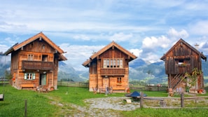 Three rustic wooden cabins are set against a backdrop of majestic mountains under a partly cloudy sky. These cabins are surrounded by lush green grass and a wooden fence. The windows of the cabins are adorned with colorful flowers, and there are several chairs outside, suggesting a cozy and inviting atmosphere.
