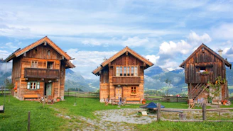 Three rustic wooden cabins are set against a backdrop of majestic mountains under a partly cloudy sky. These cabins are surrounded by lush green grass and a wooden fence. The windows of the cabins are adorned with colorful flowers, and there are several chairs outside, suggesting a cozy and inviting atmosphere.