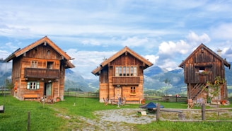 Three rustic wooden cabins are set against a backdrop of majestic mountains under a partly cloudy sky. These cabins are surrounded by lush green grass and a wooden fence. The windows of the cabins are adorned with colorful flowers, and there are several chairs outside, suggesting a cozy and inviting atmosphere.