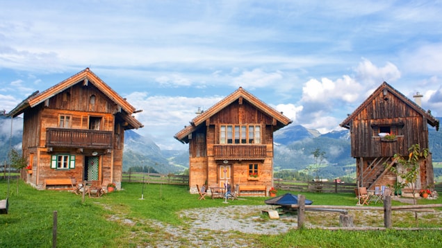 Three rustic wooden cabins are set against a backdrop of majestic mountains under a partly cloudy sky. These cabins are surrounded by lush green grass and a wooden fence. The windows of the cabins are adorned with colorful flowers, and there are several chairs outside, suggesting a cozy and inviting atmosphere.