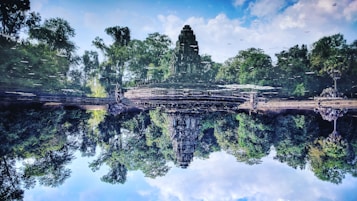 A serene landscape captures a reflection of ancient temple ruins surrounded by lush greenery in calm water. The reflection creates a symmetrical view, with vibrant green trees and a partly cloudy sky enhancing the scene.