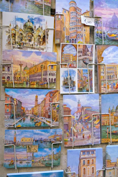 A collection of vibrant postcards showcasing various iconic scenes from Venice, including canals, gondolas, and historical architecture. The postcards are arranged in a display with some secured by metal clips. The artwork features detailed and colorful depictions of Venetian buildings and bustling streets.