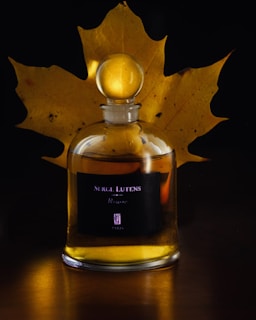 A perfume bottle labeled 'Serge Lutens' is placed in front of a large, dried maple leaf. The transparent glass of the bottle reveals a golden liquid inside, complementing the warm, autumnal hues of the leaf. The background is dark, highlighting the bottle and leaf combination.