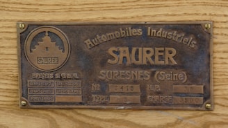 A vintage metal plate attached to a wooden surface, featuring the text 'Automobiles Industriels SAURER SURESNES (Seine)' along with patent numbers, vehicle type, horsepower, and charge weight. The plate has a textured background with a circular emblem on the left side.