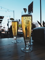 Two tall glasses of beer with lime wedges are placed on a wooden table. The setting appears to be outdoors, with string lights and a blue sky in the background. The glass is labeled with 'Craft Coast' and a logo. There is a table number visible in the distance.