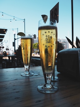 Two tall glasses of beer with lime wedges are placed on a wooden table. The setting appears to be outdoors, with string lights and a blue sky in the background. The glass is labeled with 'Craft Coast' and a logo. There is a table number visible in the distance.