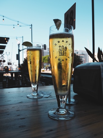Two tall glasses of beer with lime wedges are placed on a wooden table. The setting appears to be outdoors, with string lights and a blue sky in the background. The glass is labeled with 'Craft Coast' and a logo. There is a table number visible in the distance.