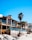A row of modern beachfront houses with large glass windows and balconies, featuring a mix of stone and wood exteriors. Palm trees are positioned along the sidewalk adjacent to the sandy beach, creating a laid-back coastal vibe. The clear blue sky suggests a sunny day.