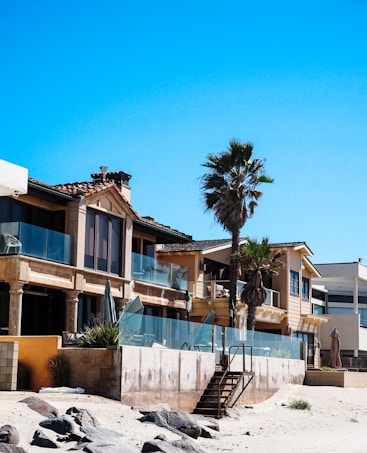 A row of modern beachfront houses with large glass windows and balconies, featuring a mix of stone and wood exteriors. Palm trees are positioned along the sidewalk adjacent to the sandy beach, creating a laid-back coastal vibe. The clear blue sky suggests a sunny day.