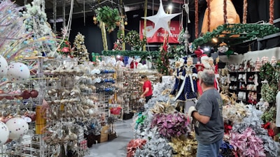 A festive store display heavily decorated with Christmas ornaments, including trees adorned with lights, garlands, and baubles. Large Santa Claus figures and mannequins in blue robes are prominently featured, surrounded by sparkling decorations in gold, silver, pink, and other hues. The ceiling is decorated with hanging greenery and a large white star.