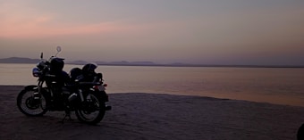 A motorcycle parked on a sandy beach with a serene water body in the background, silhouetted against a pastel-colored sunset. Distant hills are visible across the water under a calm evening sky.