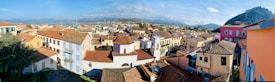 A panoramic view of a picturesque town with a mix of colorful residential buildings, featuring red-tiled roofs. The scene is framed by a mountainous backdrop and a bright, clear sky. In the distance, a large body of water is visible, giving the town a serene and idyllic atmosphere.