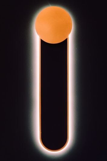 A minimalistic design featuring a vertical black bar with a rounded top, outlined by a soft glow. The top of the bar is capped with a glowing orange circle, creating a striking contrast against the dark background.