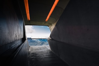 A modern interior passageway leads to a bright view of mountains under a blue sky. The smooth, curved walls and ceiling exhibit a minimalist architectural design, highlighted by orange lighting strips on the ceiling.