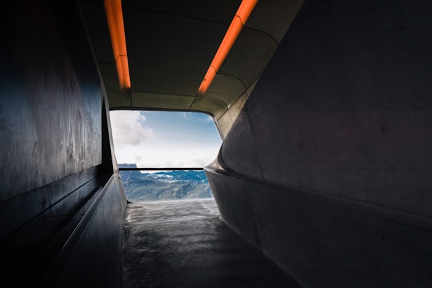 A modern interior passageway leads to a bright view of mountains under a blue sky. The smooth, curved walls and ceiling exhibit a minimalist architectural design, highlighted by orange lighting strips on the ceiling.