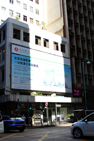 A multi-story urban building with a prominent billboard in a mix of white and light blue colors, partially illuminated by sunlight. The street below shows parked cars, a pedestrian, a small kiosk, and a bicycle. Shadows cover parts of the building, creating a contrast against the bright sections.