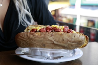 A large baked potato is placed on a white plate, topped with chopped sausages and corn. The potato skin appears crispy and brown. The dish is resting on a disposable take-out container. A person with blonde hair is partially visible in the background, wearing a dark top.