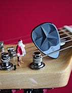 A close-up of a guitar headstock with strings and tuning pegs, featuring a small figurine of Santa Claus standing next to a guitar pick on one of the strings. The background is a solid, vibrant red.