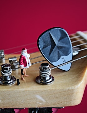 A close-up of a guitar headstock with strings and tuning pegs, featuring a small figurine of Santa Claus standing next to a guitar pick on one of the strings. The background is a solid, vibrant red.