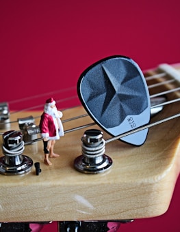 A close-up of a guitar headstock with strings and tuning pegs, featuring a small figurine of Santa Claus standing next to a guitar pick on one of the strings. The background is a solid, vibrant red.