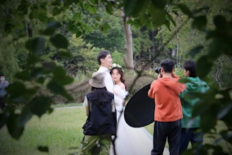 A couple is being photographed outdoors in a lush, green setting. The woman is wearing a white dress and a flower crown, while the man is dressed in light clothing. A photographer and an assistant holding a reflector are positioned in front of them. The scene is framed by foliage in the foreground.