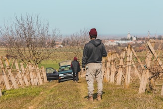 Two people dressed in casual outdoor clothing stand on a vineyard hill. One person faces the camera while the other stands near an open car door. The background reveals an expansive rural landscape with distant buildings and a clear sky.