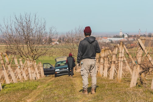 Two people dressed in casual outdoor clothing stand on a vineyard hill. One person faces the camera while the other stands near an open car door. The background reveals an expansive rural landscape with distant buildings and a clear sky.