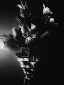 A black and white composition featuring a woven basket filled with a variety of flowers and leaves. The arrangement includes prominently visible lilies and smaller blooms against a dark background, with dramatic lighting casting shadows and highlighting textures.