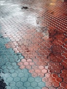 Wet hexagonal red and gray tiles with reflections from surrounding lights. Puddles of water create a glossy texture across the surface.