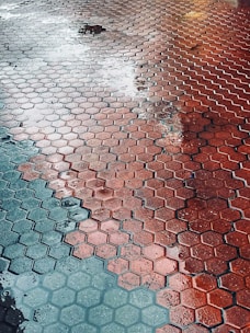 Wet hexagonal red and gray tiles with reflections from surrounding lights. Puddles of water create a glossy texture across the surface.