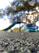 A playground structure is visible in the background, with a focus on a textured rubber surface in the foreground. The playground equipment includes a slide and is surrounded by greenery, indicated by the blurred trees.