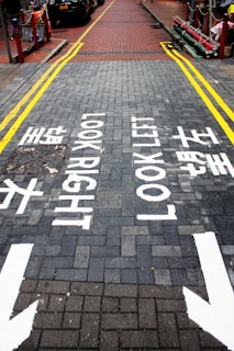 A brick-paved street with directional markings instructing pedestrians to 'Look Right' and 'Look Left' in both English and Chinese. Yellow lines border the sides of the street, and there are arrows pointing in both directions. Cars are visible parked along one side of the road.