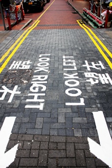 A brick-paved street with directional markings instructing pedestrians to 'Look Right' and 'Look Left' in both English and Chinese. Yellow lines border the sides of the street, and there are arrows pointing in both directions. Cars are visible parked along one side of the road.