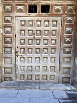 A wooden door featuring an intricate pattern of square panels, each with a carved design. The weathered texture and faded paint suggest age and history. Above the door, there are small openings with metal bars, adding a rustic touch.