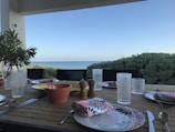 A dining table setup with plates, glasses, and cutlery is positioned on a terrace overlooking the sea. A potted plant adds greenery to the table, and a pepper grinder and napkins with floral patterns add detail. The background features a clear blue sky and lush greenery.