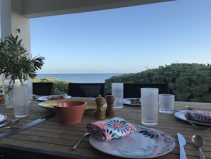 A dining table setup with plates, glasses, and cutlery is positioned on a terrace overlooking the sea. A potted plant adds greenery to the table, and a pepper grinder and napkins with floral patterns add detail. The background features a clear blue sky and lush greenery.