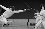 Two fencers dressed in full protective gear are engaged in a dynamic bout. One fencer is lunging forward with their épée, aiming at their opponent, while the other is in a defensive stance. The scene occurs in an indoor fencing arena, with blurred spectators and a third fencer in the background.