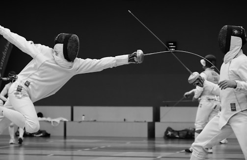 Two fencers dressed in full protective gear are engaged in a dynamic bout. One fencer is lunging forward with their épée, aiming at their opponent, while the other is in a defensive stance. The scene occurs in an indoor fencing arena, with blurred spectators and a third fencer in the background.