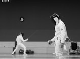 Two fencers in protective gear and helmets engage in a bout on a polished indoor floor. One fencer is in a mid-action pose with their sword extended, while the other stands in the foreground, appearing ready to defend or attack. A wheelchair is visible in the background, indicating an inclusive or adaptive fencing event. The scene is captured in black and white.
