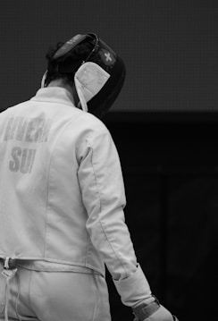 A fencer wearing protective gear, including a helmet and padded clothing, stands with their back turned. The focus is on the fencer's upper body and helmet.
