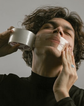 A person with curly hair is holding a roll of clear tape against their face, appearing to be stuck or wrapping it around their head. The person's eyes are closed, and they are wearing a black top. One hand has a ring on it. The background is plain gray.