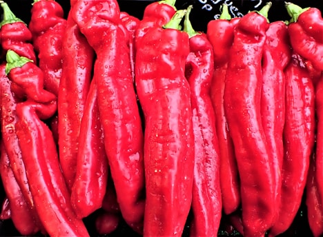 A bunch of vibrant red peppers with smooth, glossy skin and green stems. The peppers are closely arranged, filling the frame with their bright color and texture.