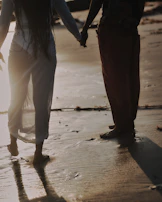 A candid shot of a couple holding hands, walking barefoot along a shoreline with charcoal shadows.
